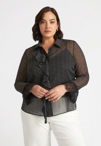 Blusa nera trasparente con pois bianchi, caratterizzata da una parte anteriore arricciata, colletto con bottoni e dettagli in pizzo sulle maniche. Si annoda in vita.