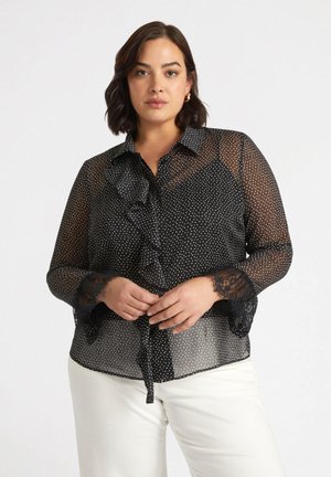 Blouse noire transparente à pois blancs, avec un devant volanté, un col à boutons et des détails en dentelle sur les manches. Se noue à la taille.