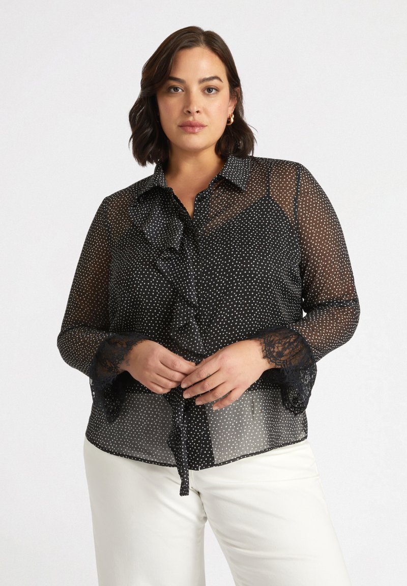 Blusa nera trasparente con pois bianchi, caratterizzata da una parte anteriore arricciata, colletto con bottoni e dettagli in pizzo sulle maniche. Si annoda in vita.