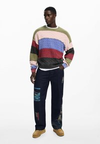 Multicolored knitted sweater with textured stripes; denim pants featuring fabric patches; brown shoes.