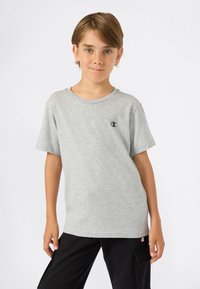 Jong persoon met lichtbruin haar, gekleed in een grijze Champion t-shirt en zwarte cargo broek, staande tegen een effen witte achtergrond.