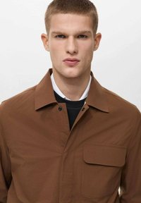Brown button-up jacket with a collar, single chest pocket, and smooth, matte texture. Worn over a black top, minimalistic design.