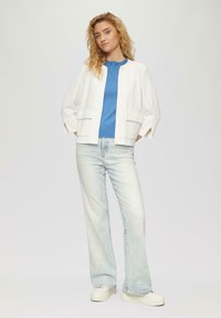 Veste blanche cropped avec deux poches avant, associée à un pull bleu à col rond et à un jean flare bleu clair. Baskets en toile.