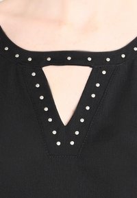 Svart topp med V-ringning och utskärning, utsmyckad med silverfärgade nitar runt halsringningen, texturen verkar slät och matt.