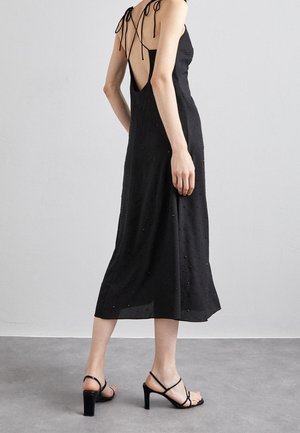 Vrouw in een zwarte mouwloze midi-jurk met een open rug en gestrikte bandjes, draagt zwarte sandalen met brede hak en bandjes, op een grijze vloer.