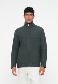 Casaco de polar verde escuro com gola alta, fecho frontal com zip e punhos canelados. Apresenta um acabamento texturizado e detalhe de costura horizontal.