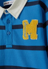 Modrá pruhovaná polokošeľa s bielym golierom, modrými gombíkmi a žltou chlpatou nášivkou "M" s modrým šitím. Vyrobená z kombinácie bavlny.