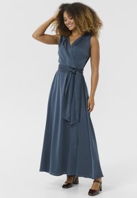 Robe portefeuille en tissu bleu marine, sans manches avec un décolleté en V profond, nouée à la taille. Texture lisse et jupe longue. Chaussures à talons carrés marron.