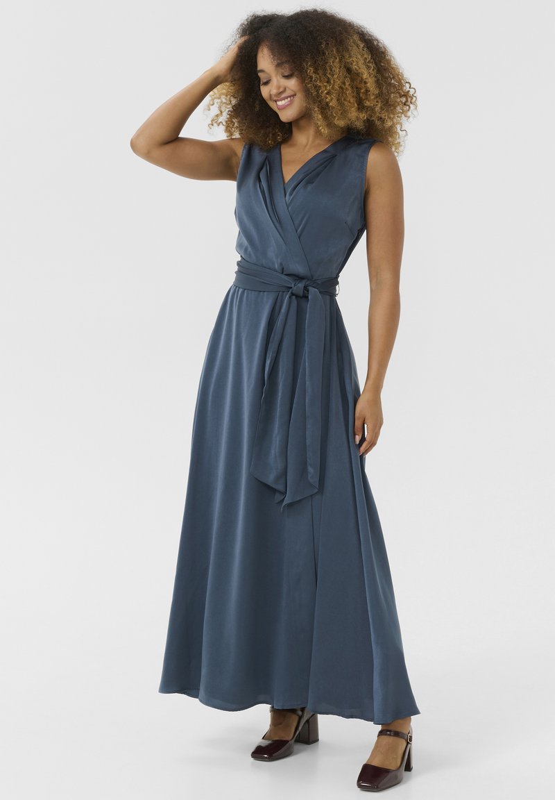 Robe portefeuille en tissu bleu marine, sans manches avec un décolleté en V profond, nouée à la taille. Texture lisse et jupe longue. Chaussures à talons carrés marron.