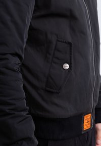 Giacca bomber nera con chiusura a zip, tessuto strutturato, tasca laterale con bottone metallico e etichetta arancione sulla vita.
