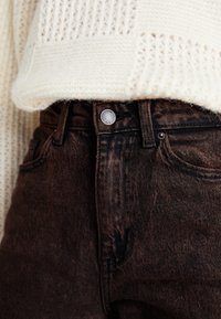Gros plan sur la taille d'un jean en denim marron foncé et le bouton avant, associé à un pull en tricot écru partiellement rentré.