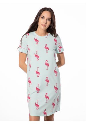 Frau trägt hellblaues kurzärmeliges Kleid mit pinkem Flamingo-Print, steht mit den Händen hinter dem Rücken vor einem weißen Hintergrund.