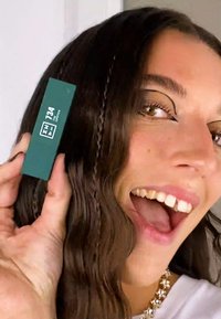 Grønn rektangulær produkt merket "ZH 734 Impact" holdt i en hånd, med naturlig bølgete hår og en noe uskarp bakgrunn.