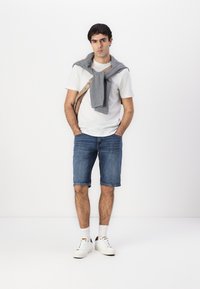 T-shirt blanc à manches courtes, shorts en denim bleu, sweat gris noué autour du cou, sac à bandoulière beige, sneakers blancs avec des accents noirs.