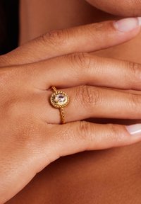 Gouden ring met een gedraaide band, voorzien van een ovale steen omringd door een gestructureerd frame, gezet op een hand met een lichte huidtoon.