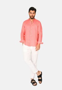 Camicia button-up di lino corallo con maniche lunghe, abbinata a pantaloni di lino bianchi e sandali neri, in piedi su uno sfondo semplice.
