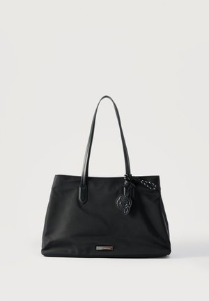 Borsa tote nera con manici sottili, piastra metallica con marchio e ciondolo decorativo in pelle appeso a una delle cinghie, posata su uno sfondo bianco uniforme.