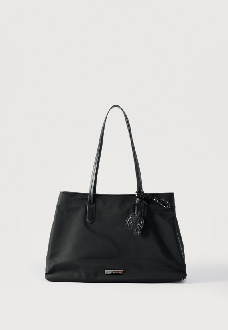 Μαύρη τσάντα χειρός με λεπτές λαβές, μεταλλική πλακέτα με λογότυπο και διακοσμητικό δερμάτινο γούρι που κρέμεται από ένα λουρί, τοποθετημένη σε απλό λευκό φόντο.