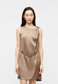 Γυναίκα με μακριά σκούρα μαλλιά, φορώντας αμάνικο, γυαλιστερό φόρεμα σε χρώμα ταουπέ με ασύμμετρο ντεκολτέ και λεπτή μεταλλική αλυσίδα στη μέση, στέκεται μπροστά σε λευκό φόντο.