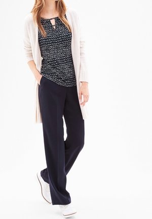 Frau trägt eine hellbeige Strickjacke, ein schwarz-weiß gemustertes Oberteil, schwarze weit geschnittene Hose und weiße Turnschuhe, steht mit einer Hand in der Tasche.