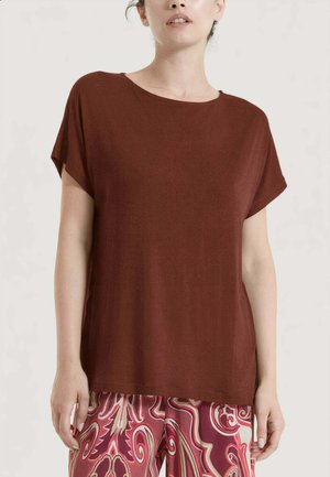 Donna con una maglietta marrone a maniche corte larga e pantaloni rossi con fantasia a gamba larga, in piedi davanti a uno sfondo chiaro e semplice.