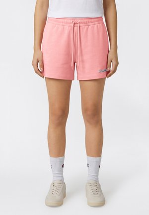 Kobieta stojąca, ubrana w różowe szorty z troczkiem, białe skarpetki do kostek z logo, białe sneakersy oraz biały top, na jednolitym jasnym tle.