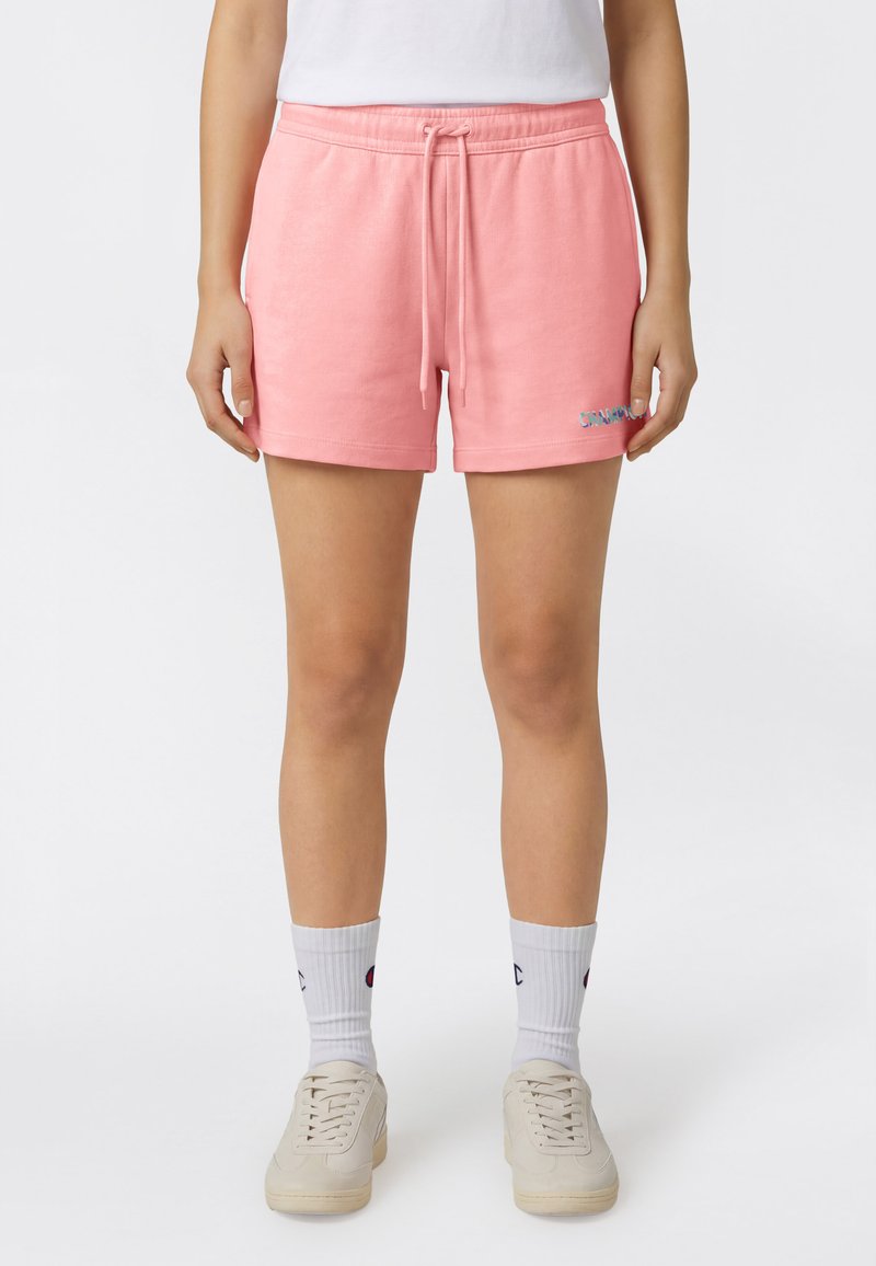 Femme debout portant un short rose à cordon de serrage, des chaussettes blanches montantes avec logos, des baskets blanches et un haut blanc, sur un fond clair uni.
