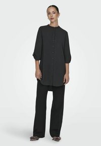 Zwarte blouse met een ronde kraag, knoopsluiting aan de voorkant en driekwartmouwen, gecombineerd met zwarte wijde broek en schoenen met luipaardprint.