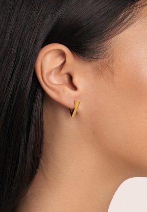 Orecchio di donna con capelli scuri e lisci, che indossa un piccolo orecchino a cerchio geometricamente a forma di V in oro su uno sfondo semplice.