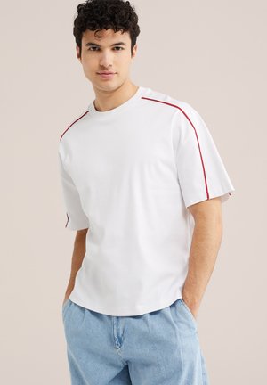 Młody mężczyzna z kręconymi włosami, ubrany w białą koszulę z krótkim rękawem i czerwonymi paskami na ramionach oraz jasnoniebieskie dżinsy, ręce w kieszeniach, neutralne tło.