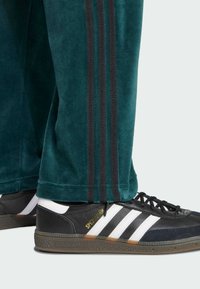 Zapatilla Adidas Spezial en blanco y negro, combinada con pantalones de chándal en color teal que presentan tres franjas negras a lo largo de la pierna.
