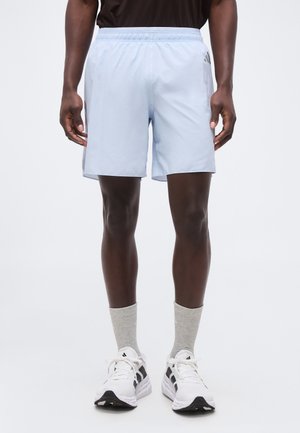 Mann trägt hellblaue Adidas-Sportshorts, graue Socken, weiße Adidas-Sneaker und steht vor einem schlichten Hintergrund.