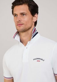 Camisa polo blanca con cuello alzado que presenta rayas rojas y azul marino. Logos bordados en el pecho. Corte ajustado con mangas cortas.