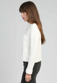 Femme avec de longs cheveux bruns portant un pull blanc à manches longues et un jean gris foncé, tournée vers la gauche devant un fond gris uni.