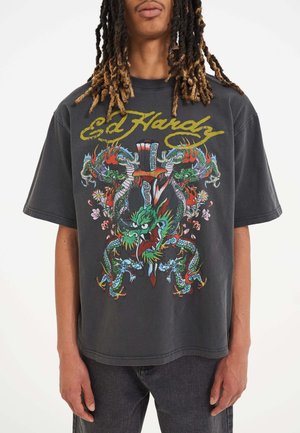 Camiseta de gran tamaño en gris con gráficos vibrantes de dragones y elementos florales, con el texto "Ed Hardy" en amarillo en la parte superior. Diseño casual.