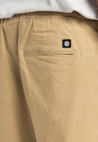 Beige bomuldsshorts med elastisk talje, baglomme med en horisontal søm og et sort logo-patch på lommen.
