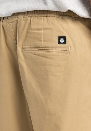 Beige cotton shorts featuring an elastic waistband, back pocket with a horizontal seam, and a black logo patch on the pocket.
