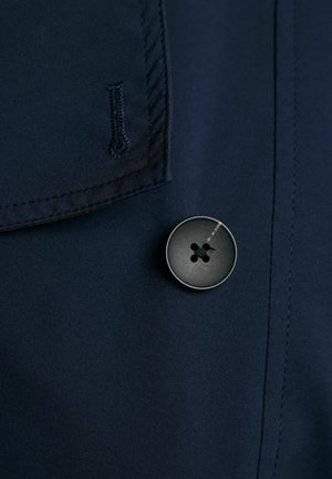 Detal niebieskiego materiału z dużym, ciemnym guzikiem z czterema otworami, w towarzystwie przeszytej krawędzi kieszeni i gładkiej tekstury.