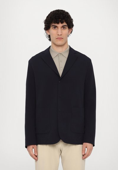 Blazer bleu marine en tissu doux et texturé, avec un design à boutonnage simple, deux poches avant et un col cranté sur une chemise beige.