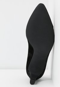 Semelle de chaussure noire à bout pointu avec un petit talon, présentée sur un fond blanc et un mur.