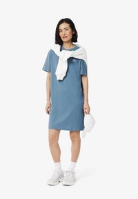 Abito a T-shirt in cotone blu con maniche corte, caratterizzato da un logo ricamato. Maglione bianco annodato attorno alle spalle, abbinato a sneaker bianche.