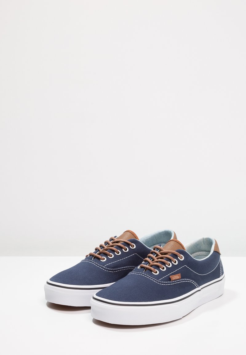 Un par de zapatillas Vans de lona azul marino con detalles de cuero marrón y suelas de goma blanca sobre una superficie blanca frente a un fondo plano.