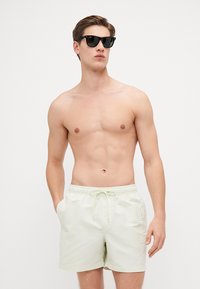 Shorts de bain vert clair en tissu lisse, dotés d'une taille élastique avec cordon de serrage et d'une poche latérale. Texte du logo sur la jambe.