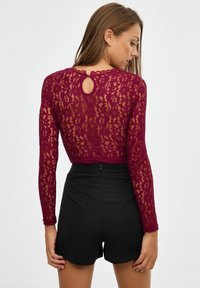 Top de encaje burdeos con mangas largas, que presenta un escote en forma de lágrima en la parte posterior y un estampado floral, combinado con pantalones cortos negros de cintura alta.