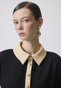 Zwarte jurk met een crèmekleurige kraag en vijf decoratieve gouden knopen aan de voorkant. Model draagt gouden druppeloorbellen. Gladde textuur.