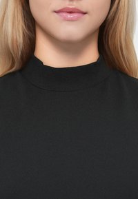 Svart høyhalset topp laget av glatt stoff. Har en tettsittende krage og et minimalistisk design, uten synlige mønstre eller dekorasjoner.