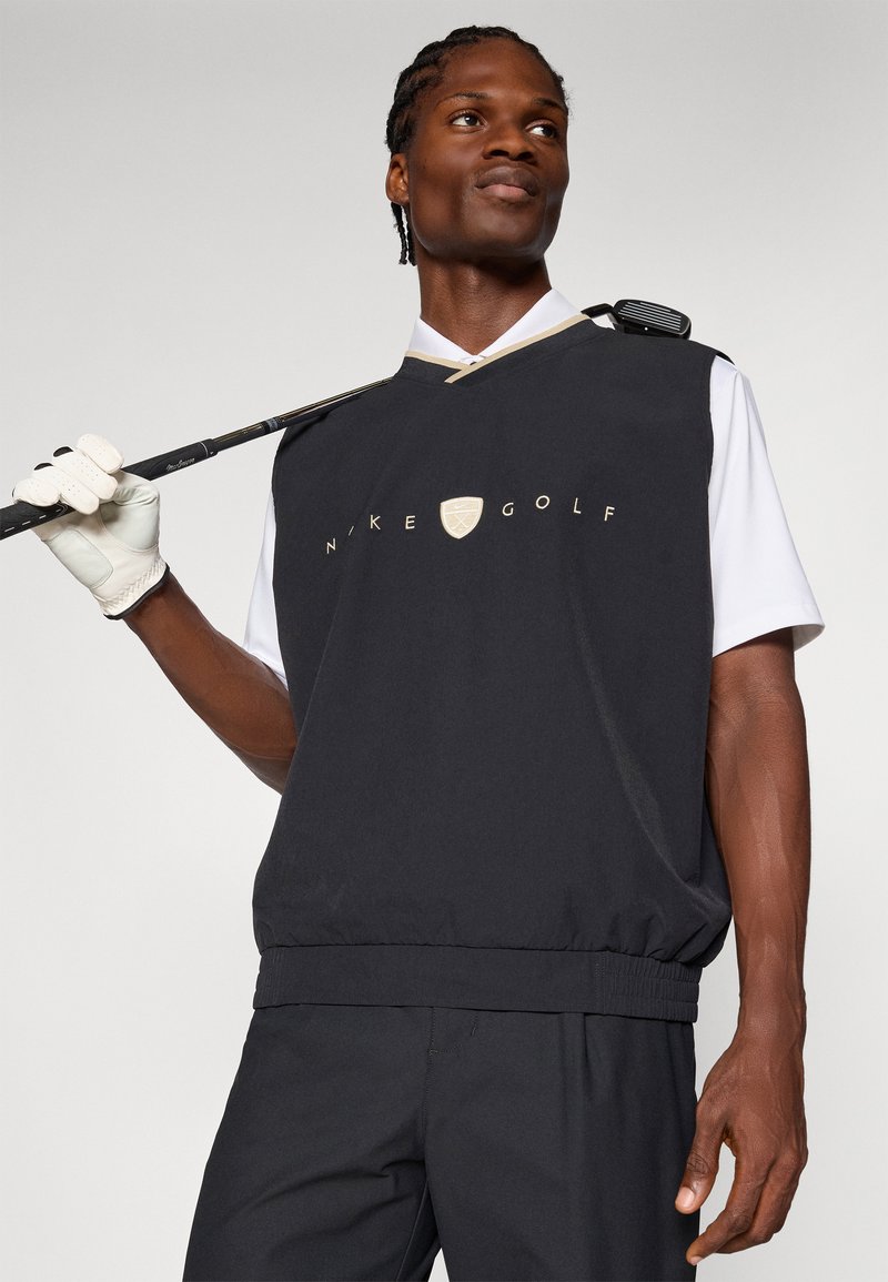Gilet de golf noir Nike avec un logo doré, présentant un col ras du cou, une taille élastique et une chemise blanche empilée en dessous, tenue par un golfeur.