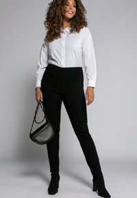 Camicia bianca con bottoni, pantaloni neri su misura e stivaletti con tacco. La modella tiene una borsa a tracolla nera. Tessuto liscio con un design aderente.