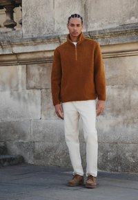 Brauner Fleece-Pullover mit einem Viertel-Reißverschluss, kombiniert mit weißen Hosen und braunen Schnürstiefeln. Weiche Textur mit entspannter Passform.