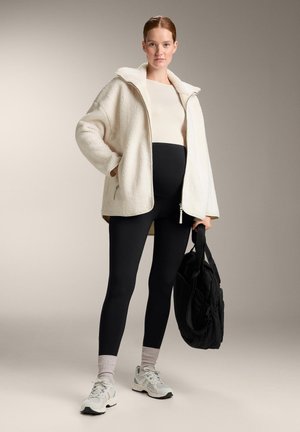 Bela jopa z zadrgo iz flisa z visokim ovratnikom, črne legice, sive superge in črna torbica. Model ima sproščen položaj.